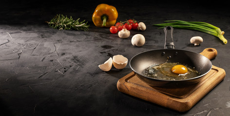 Chicken egg in empty pan surrounded by vegetarian cuisine ingredients on dark table