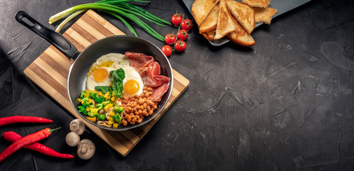 Healthy breakfast with fried eggs, bacon, beans, toasts, mushrooms, broccoli and tomatoes on black background. Top view