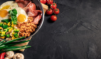 Healthy breakfast with fried eggs, bacon, beans, toasts, mushrooms, broccoli and tomatoes on black background. Top view