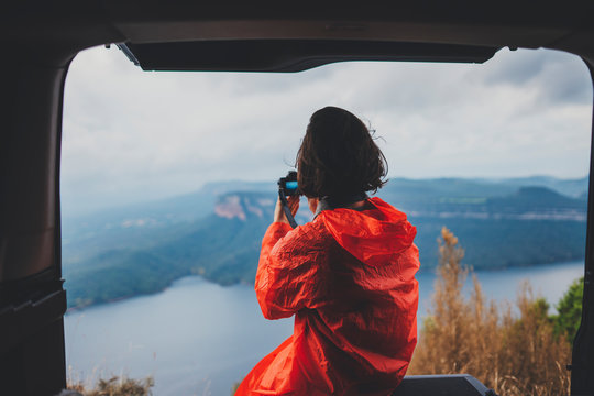 Back view of young traveller hipster girl sitting in trunk of car and taking photo of amazing nature landscape, young woman exploring hiking routes in mountains, Discover Explore Enjoy Travel Concept