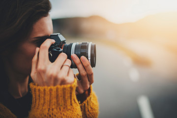 Closeup of young hipster girl taking photograph on vintage camera while standing on road capturing amazing nature landscape, traveller woman in cozy jersey enjoying vacation in Europe - obrazy, fototapety, plakaty