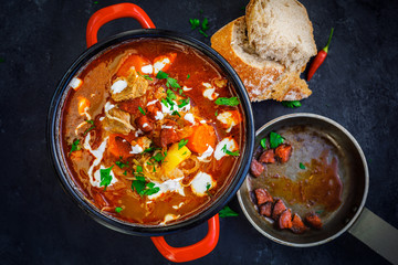 goulash soup in red pot and bread slices on rustic black board background - obrazy, fototapety, plakaty