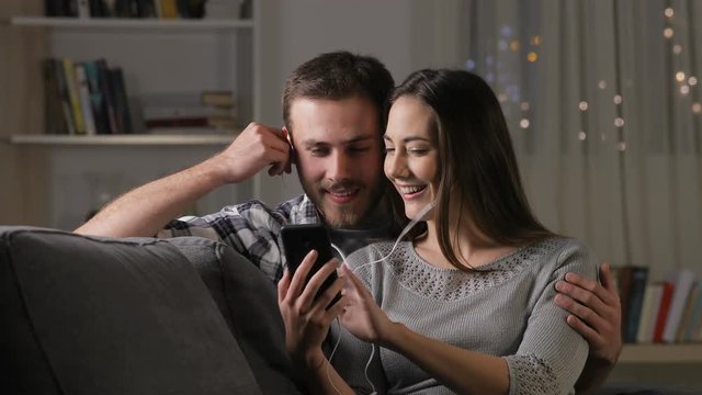 Happy couple sharing earphones watching videos online on a smart phone sitting on a couch at home in the night