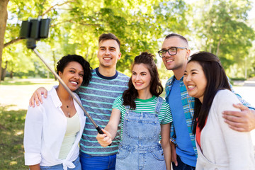 people, technology, friendship and international concept - group of happy friends or students taking picture by smartpnone on selfie stick at summer park