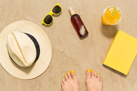 vacation and summer holidays concept - female feet, sunglasses, straw hat, sunscreen and juice with book on beach sand