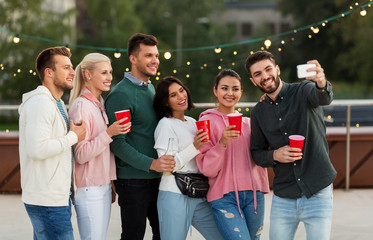 leisure and people concept - happy friends with drinks in party cups taking selfie by smartphone on rooftop in summer