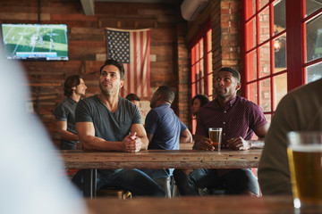 Male Friends Drinking Beer And Watching Game On Screen In Sports Bar