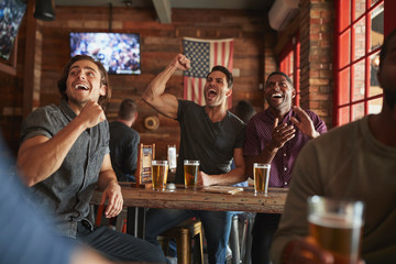 Group Of Male Friends Celebrating Whilst Watching Game On Screen In Sports Bar