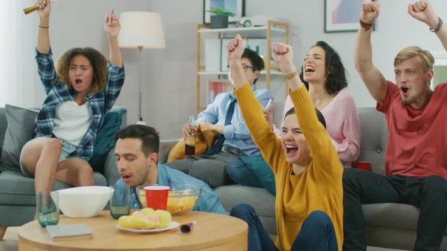 At Home Diverse Group of Sports Fans Wearing Team's Uniform Watch Sports Game Match on TV, They Cheer and Chant for the Team, Celebrate Victory after Team Scores a Winning Goal. Cozy Room with Snacks