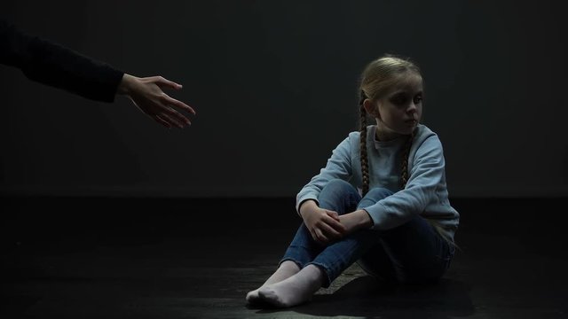 Distrustful little girl sitting in dark room refusing adult helping hand, fear