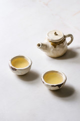 Hot green tea in two traditional chinese clay ceramic cup and teapot standing on white marble table.