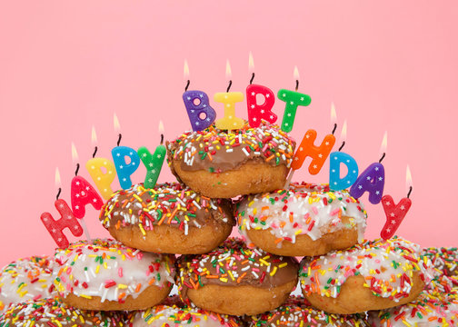 Chocolate and white frosted donuts covered in candy sprinkles piled into a cake pile with Happy Birthday candles burning brightly. Pink background.