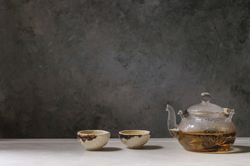 Hot green tea in two traditional chinese clay ceramic cup and glass teapot standing on white marble table.