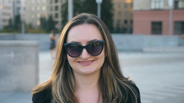 Portrait of young businesswoman in sunglasses with cityscape at background. Happy attractive business woman looking at the camera and straightens hair. Face of smiling confident girl. Close up