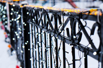 Metal forged fence is decorated with garlands, multi-colored lights, a beautiful blurred background.
