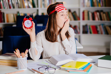 Tired sleepy teenager student woman yawning while reparing for exams late night at library. Young college girl in sleep mask working on desk with notebooks, books and notes. Tired lady study overtime.