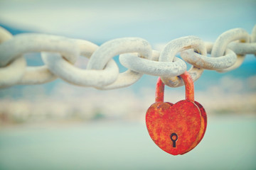 Vintage red heart shaped padlock as symbol of eternal love hanging on chain link at dock with sea...