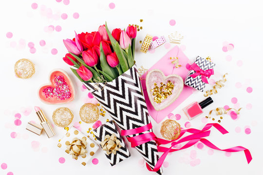 Pink and Gold Styled Desk with Florals. Pink tulips in black and white stylish wrapping paper, gifts, cosmetics and female accessories with confetti on white background