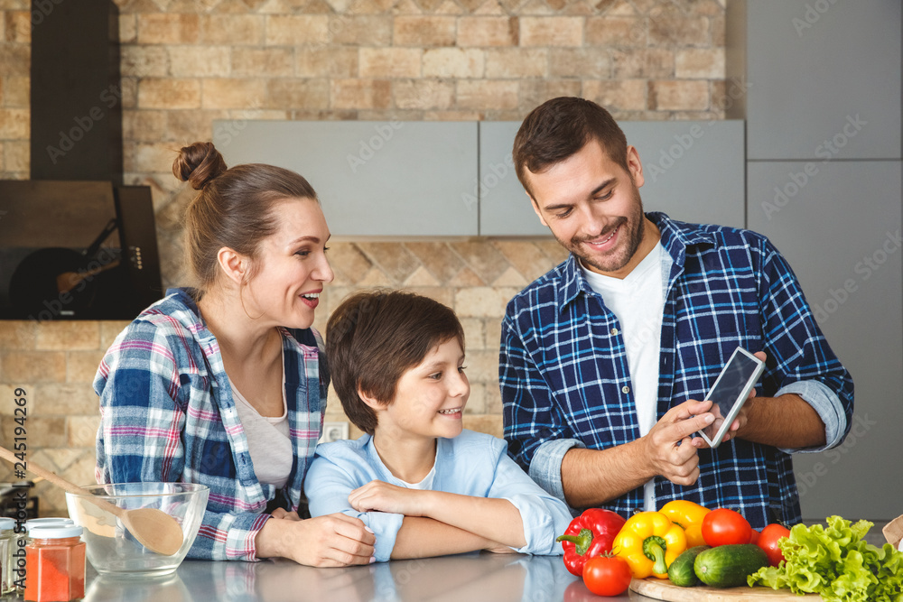 Wall mural Family at home standing in kitchen together mother and son smiling curious looking at father showing app on digital tablet