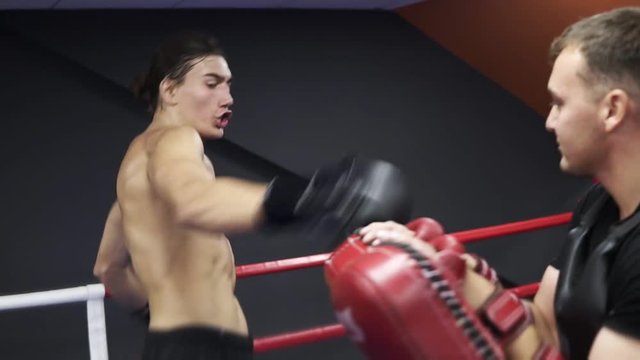 Caucasian man shirtless in black shirts training at gym. Young msn train his legs by working with paws, hitting. Boxing ring