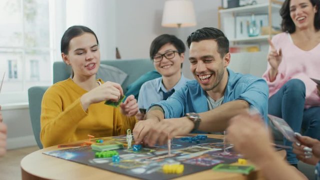 Diverse Group of Guys and Girls Playing in a Strategic Board Game with Cards and Dice. Team Players Do High Five. Cozy Living Room in a Daytime