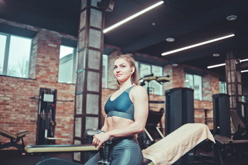 Fototapeta na wymiar Training pause, sporty young woman in sportswear resting on bench holding dumbbells in her hands and listens to music in headphones at gym