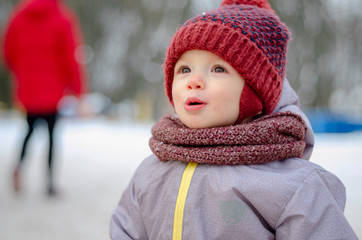 Portrait of a cute baby dressed in a gray jacket and a red hat that walks through the snow covered snow park. She smiles one in the photo during the snowfall