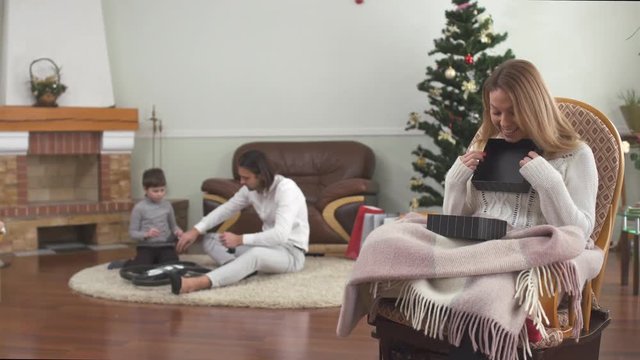Happy young mother sitting in an armchair opening a box pleasantly surprised by a gift. Father and little son playing with drone sitting on the floor in the background. Family celebrates xmas at home.