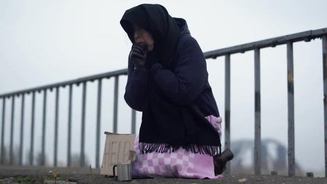 Adult homeless woman sits on the bridge in cold windy grey weather asking for alms and help and coughing