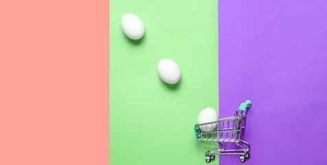 Chicken eggs in mini shopping trolley for shopping on colored background, consumer concept, minimalism, top view..