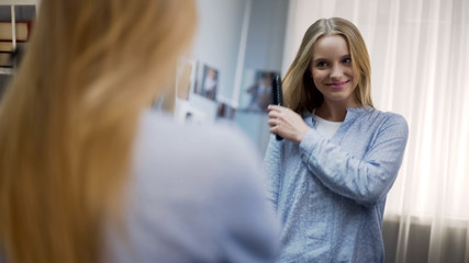 Haircare treatment, smiling beautiful female combing long blond hair near mirror