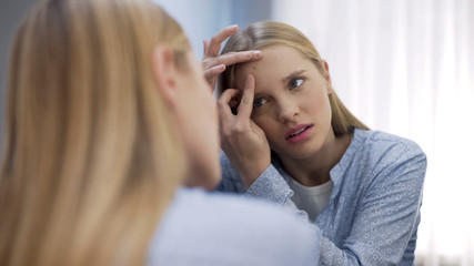 Worried young woman squishing pimples on face in front of mirror, insecurities