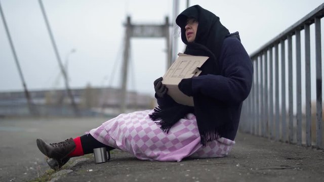 Adult homeless woman with carton sits on the bridge in cold windy grey weather asking for alms and help