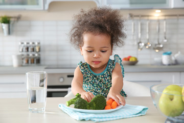 Cute African-American girl eating vegetables at table in kitchen