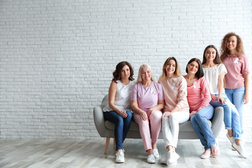 Group of women with silk ribbons on sofa against brick wall, space for text. Breast cancer...