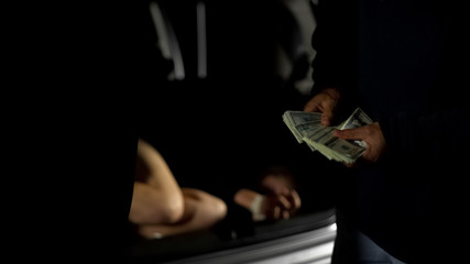 Male criminal paying kidnapper for tied female victim lying in auto trunk