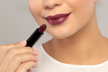 Beautiful woman applying dark lipstick on gray background, closeup