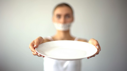 Skinny woman with taped mouth showing empty plate, concept of fasting, hunger - obrazy, fototapety, plakaty