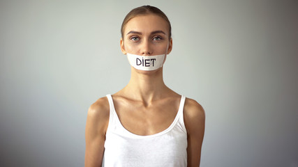 Exhausting diet concept, miserable slim woman with taped mouth looking at camera - obrazy, fototapety, plakaty