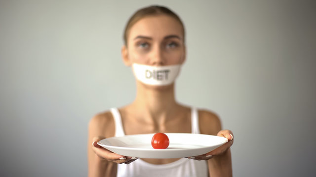 Thin girl with taped mouth holding plate with tomato, exhausting diet, anorexia