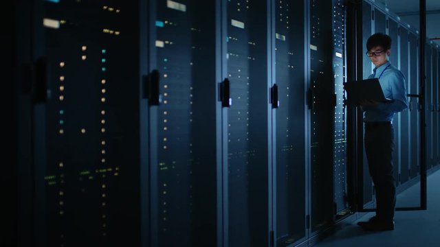 Dark Data Center: Male IT Specialist Stands Beside the Row of Operational Server Racks, Uses Laptop for Maintenance. Concept for Cloud Computing, Artificial Intelligence, Supercomputer, Cybersecurity