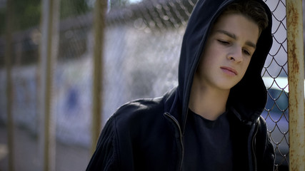Boy in hoodie leaning on fence, orphan in boarding school, confinement - obrazy, fototapety, plakaty