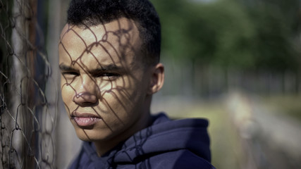 Poor afro-american teenager standing by metal fence, life difficulties concept