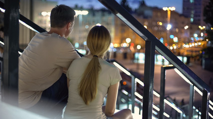 Fototapeta premium Couple sitting on stairs, embracing, watching illuminated evening city, love