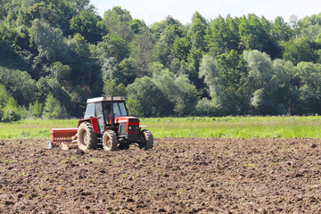 Obraz premium A farmer on a red tractor with a seeder sows grain in plowed land in a private field in the village area. Mechanization of spring field work. Farmer's everyday life. Processing of land. The agrarian.