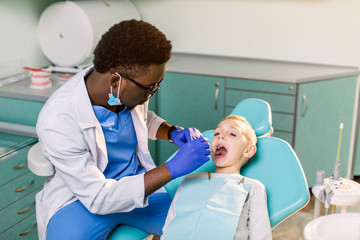 African-American ethnic black male dentist in blue latex gloves checking condition of teeth of little smiling boy. baby boy in blue dental chair in modern dentistry clinic