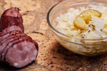 Traditional organic raw and fermented sauerkraut with pickled Persian cucumbers and homemade smoked pork sausage on olive wood cutting board.