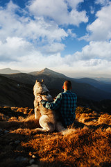 Alone tourist sitting with Alaskan Malamute dog in mountains. Man with his dog on mountain top. Hiker with dog looking at beautiful view in mountains