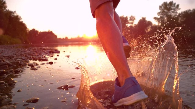 SLOW MOTION TIME REMAP CLOSE UP: Unrecognizable sportsman running in the river shallows at golden hour. Cinematic shot of male jogger splashing stream water as he runs towards the picturesque sunset.