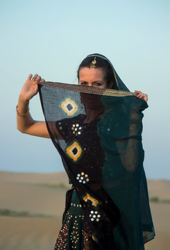 A girl in Indian clothes hides her face behind a veil against the sky and dunes. Rajasthan, India.